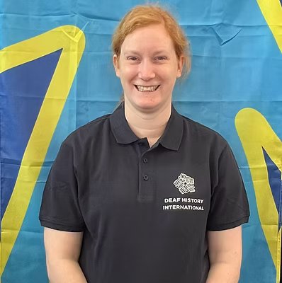 A white woman with red hair and a big smile, wearing a DHI polo shirt in front of the deaf flag.