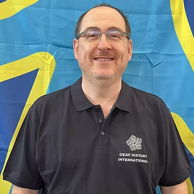 A white man wearing glasses and a DHI polo shirt in front of the deaf flag.