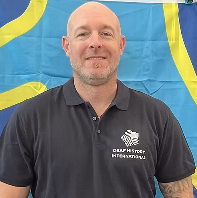 A white man with a tattoo on one arm, wearing a DHI polo shirt in front of the deaf flag.