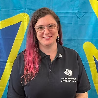 A white person with pink hair, wearing a DHI polo shirt in front of the deaf flag.