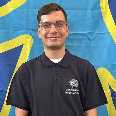 A white man with a big smile, wearing glasses and a DHI polo shirt in front of the deaf flag.