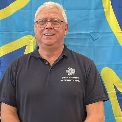 A white man with white hair, wearing glasses and a DHI polo shirt in front of the deaf flag.